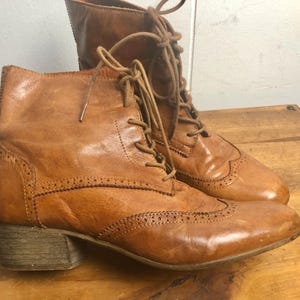 May include: A pair of brown leather lace-up boots with a brogue design. The boots have a block heel and a rounded toe.
