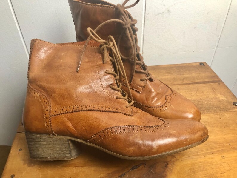 May include: A pair of brown leather lace-up boots with a brogue design. The boots have a block heel and a rounded toe.