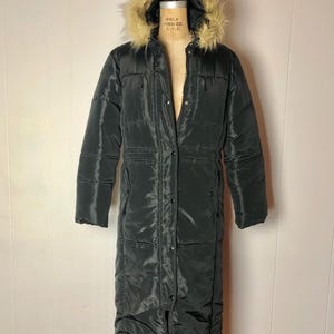 Könnte beinhalten: Ein langer, schwarzer Steppmantel mit einer mit Kunstfell gefütterten Kapuze. Der Mantel hat einen Knopf- und Reißverschluss sowie Taschen. Der Mantel wird auf einer Vintage-Schneiderpuppe präsentiert. Ein Wintermantel für kaltes Wetter.