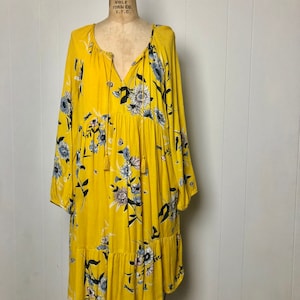 May include: A yellow dress with long sleeves and a floral pattern in blue, white, and black. The dress has a V-neckline with a tassel tie and a ruffled hem. The dress is displayed on a vintage dress form.