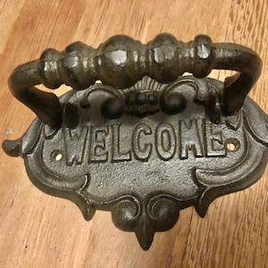 Puede incluir: Un llamador de puerta de hierro fundido negro con un asa decorativa y la palabra "WELCOME" inscrita en la parte delantera.