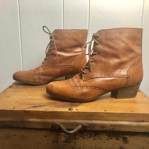 May include: A pair of brown leather ankle boots with lace-up closures and a brogue design. The boots have a low heel and are made of a soft, supple leather.