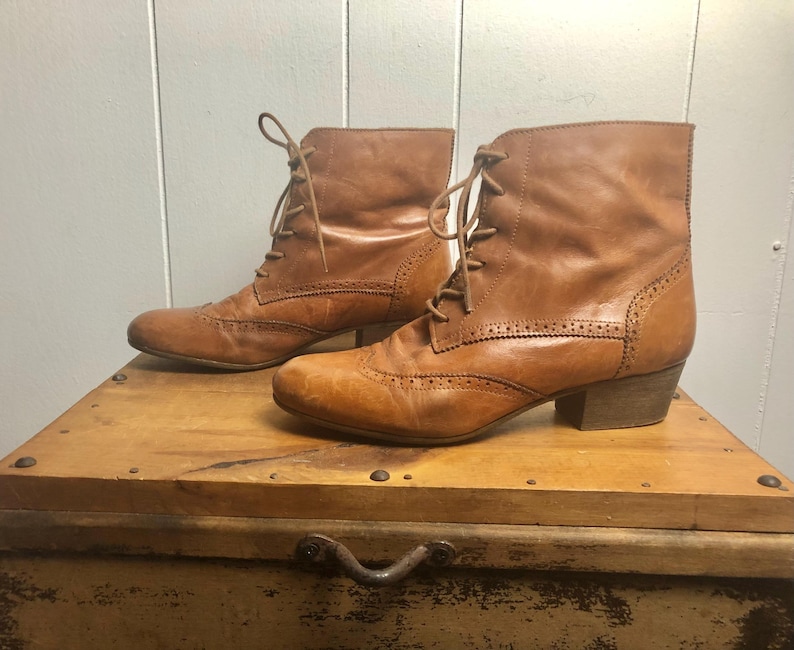 May include: A pair of brown leather ankle boots with lace-up closures and a brogue design. The boots have a low heel and are made of a soft, supple leather.