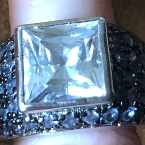 Könnte beinhalten: Ein silberner Ring mit einem großen, quadratischen, klaren Edelstein in der Mitte. Das Band ist mit kleinen, dunklen Edelsteinen besetzt. Der Ring ist ein auffälliges Schmuckstück für besondere Anlässe.