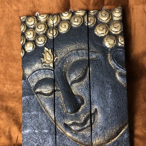 Könnte beinhalten: Eine dreiteilige Wandskulptur eines Buddha-Gesichts, aus Holz geschnitzt und in Schwarz mit goldenen Akzenten bemalt. Das Gesicht ist ruhig und die Augen sind geschlossen.