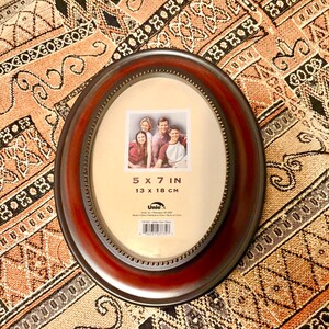 May include: An oval-shaped picture frame with a dark wood finish and a gold trim. The frame is 5x7 inches or 13x18 cm. It has a label that says "Uniquely You" and "Katelyn Dual Cherry".