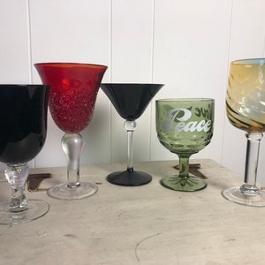 Könnte beinhalten: Fünf verschiedene Glaskelche, jeder mit einem einzigartigen Design. Die Kelche sind in einer Reihe auf einer weißen Holzoberfläche angeordnet. Die Kelche sind schwarz, rot, grün und klar mit einem braunen Wirbel.