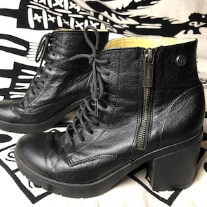 Könnte beinhalten: Ein Paar schwarze Lederstiefel mit einem dicken Absatz und Schnürung vorne. Die Stiefel haben einen seitlichen Reißverschluss und ein kleines Metall-Logo an der Seite.