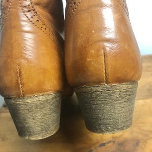 May include: A pair of brown leather boots with a brogue design. The boots have a low heel and a rounded toe.