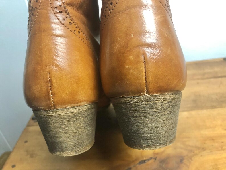 May include: A pair of brown leather boots with a brogue design. The boots have a low heel and a rounded toe.