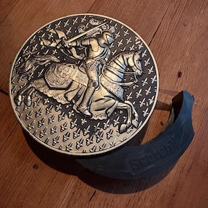 May include: A gold-colored metal plaque with a raised design of a knight on horseback. The knight is wearing armor and holding a sword. The background of the plaque is decorated with a pattern of fleur-de-lis.