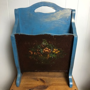 May include: A blue wooden magazine rack with a painted floral design on the front. The rack has a handle on the top and stands on two legs.