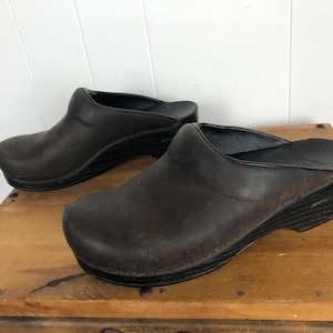 Può includere: Un paio di zoccoli in pelle marrone con suole nere. Gli zoccoli hanno un aspetto usurato e sono su una superficie di legno.