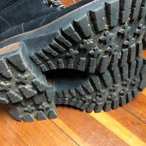 May include: Close-up of the black rubber soles of a pair of boots. The soles have a tread pattern with a series of raised ridges and cross-hatch markings.