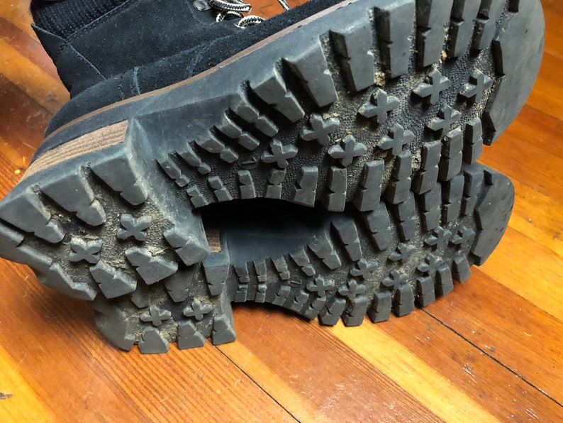 May include: Close-up of the black rubber soles of a pair of boots. The soles have a tread pattern with a series of raised ridges and cross-hatch markings.