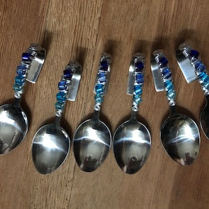 Könnte beinhalten: Sechs silberne Löffel mit blauen und weißen Glasperlen-Griffen. Die Löffel sind in einer Reihe auf einer Holzoberfläche angeordnet.