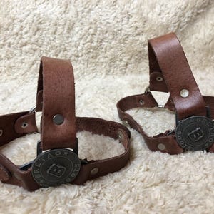 Puede incluir: Dos correas de bota de cuero marrón con hebillas de metal y un logotipo de metal que dice "Brekaox Boots".