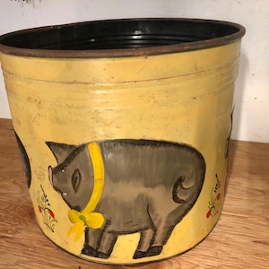 Könnte beinhalten: Ein gelber Metalltopf mit schwarzem Rand, bemalt mit einem grauen Schwein, das ein gelbes Band trägt. Das Schwein ist von grünen Blättern und roten Blumen umgeben.