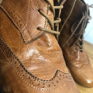 May include: Close-up of a pair of brown leather lace-up boots with brogue detailing. The boots have a worn look and are made of a high-quality leather.