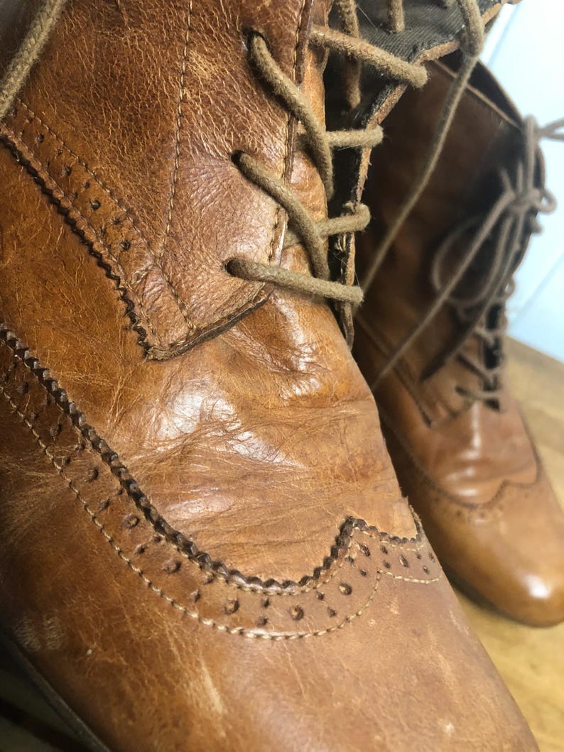 May include: Close-up of a pair of brown leather lace-up boots with brogue detailing. The boots have a worn look and are made of a high-quality leather.