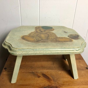 Puede incluir: Un pequeño taburete de madera pintado de verde claro con un acabado desgastado. La parte superior del taburete presenta una imagen pintada a mano de un oso de peluche con un sombrero verde. El oso está sentado y sostiene una pequeña pelota.