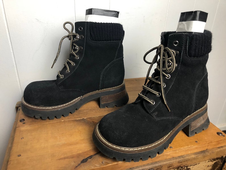 May include: A pair of black suede ankle boots with a chunky platform sole and lace-up closure. The boots have a brown trim around the sole and a black knit cuff at the top.