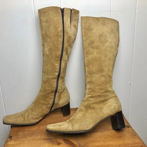 Könnte beinhalten: Ein Paar kniehohe Wildlederstiefel mit Blockabsatz. Die Stiefel sind hellbraun und haben einen Reißverschluss an der Innenseite jedes Stiefels.