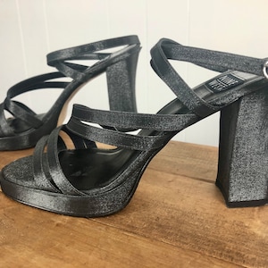 May include: A pair of black satin heeled sandals with a criss-cross strap design. The heels are block heels and the sandals have a buckle closure.