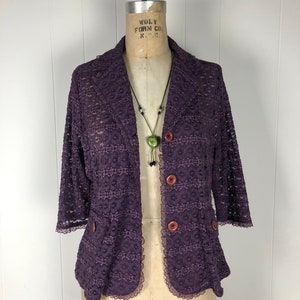 Puede incluir: Una chaqueta de encaje morado con cierre de botón y ribete de encaje. La chaqueta tiene un bolsillo en el lado izquierdo y un collar de cuentas con un colgante de corazón verde.