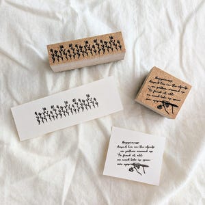 以下が含まれることがあります： 黒いインクのついた木製のスタンプが2つ。1つのスタンプには小さな花の列が描かれており、もう1つのスタンプには幸福についての引用が書かれています。引用は次のとおりです。「幸福は、それを探し出すために周囲に集める能力にあるのではなく、目を覚ます必要があるのです。」