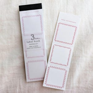 以下が含まれることがあります： ピンクの花柄の縁取りが付いた2冊の白いラベルブック。1冊は無傷で、もう1冊は開いており、3つのラベルが表示されています。無傷のブックには「3 pattern label book mini」と「Made in JAPAN」の文字があります。