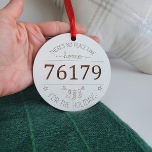 Puede incluir: Adorno navideño redondo blanco con el texto "THERE'S NO PLACE LIKE home" y "76179 FOR THE HOLIDAYS". El adorno tiene una cinta roja para colgar y presenta un diseño festivo con calcetines y estrellas.