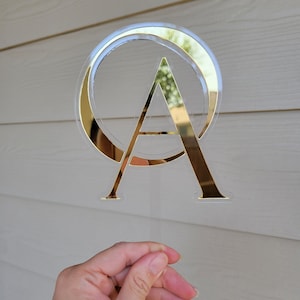 May include: A gold letter A with a gold circle around it. The letter and circle are on a clear acrylic background.