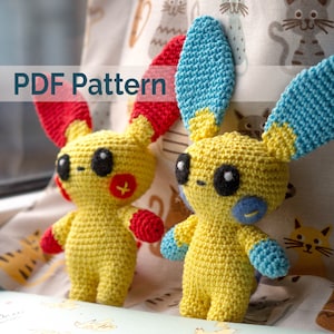 May include: PDF pattern for crocheting two yellow amigurumi plushies with blue and red accents. The plushies have large black eyes and are sitting on a fabric background with a cat pattern.