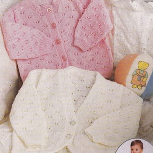 May include: Two knitted baby cardigans. One is light pink with button closures and a floral pattern. The other is off-white with a similar design. A small toy with a bear graphic is also visible.