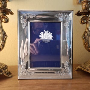 Op de afbeelding: Een rechthoekige zilverkleurige fotolijst met in reliëf aangebrachte bloemendetails in de hoeken. Het midden bevat een donkerblauw inzetstuk met een wit grafisch logo van twee cherubijnen en de tekst VITOPARDO ARGENTI in witte hoofdletters.