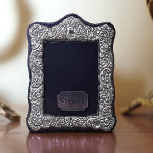 Puede incluir: Marco de fotos de plata de ley ornamentado con un respaldo de terciopelo azul oscuro. El marco presenta intrincados diseños florales y de volutas. Una pequeña etiqueta rectangular dice "Sterling Silver Made in England."