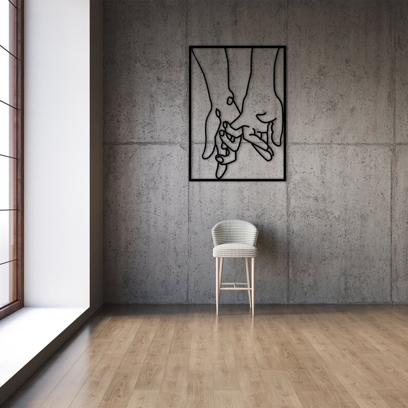 May include: A black metal wall art sculpture depicting three hands holding each other. The sculpture is framed in a black square frame and is hanging on a gray concrete wall.