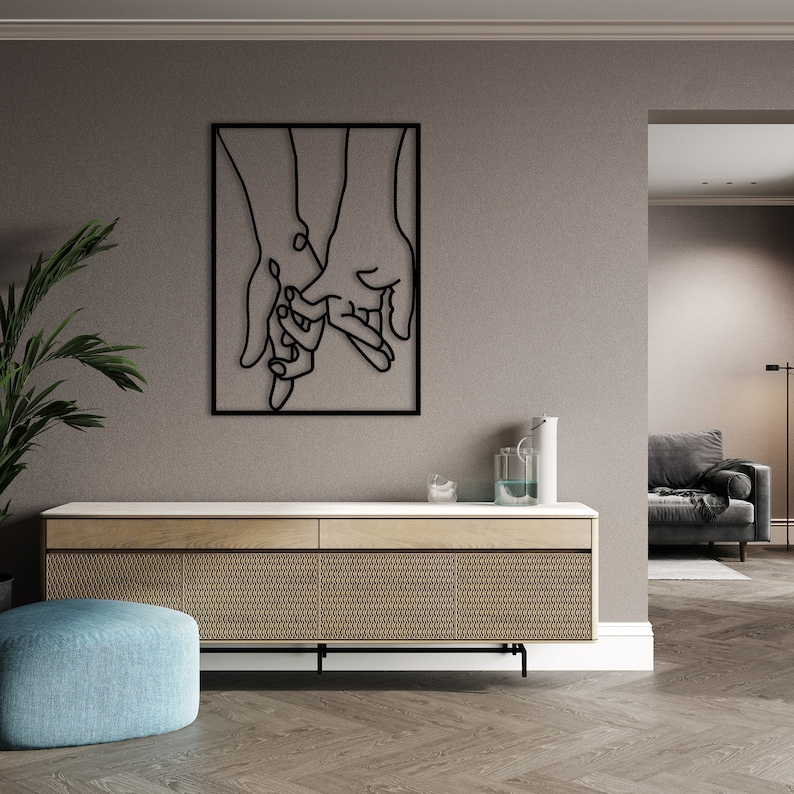 May include: Black metal wall art depicting two hands holding each other. The art is framed and mounted on a light gray wall.