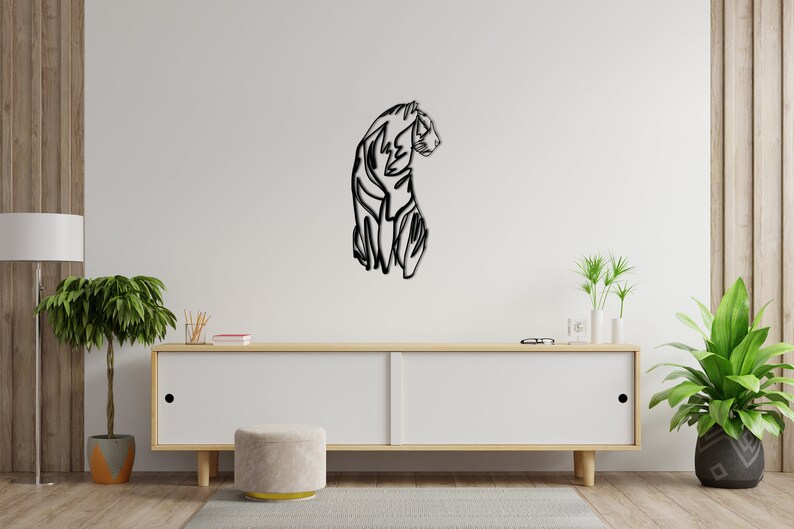 May include: A black and white line art wall decor of a tiger in a sitting pose. The tiger is facing to the right and has a long tail. The decor is mounted on a white wall above a wooden cabinet.