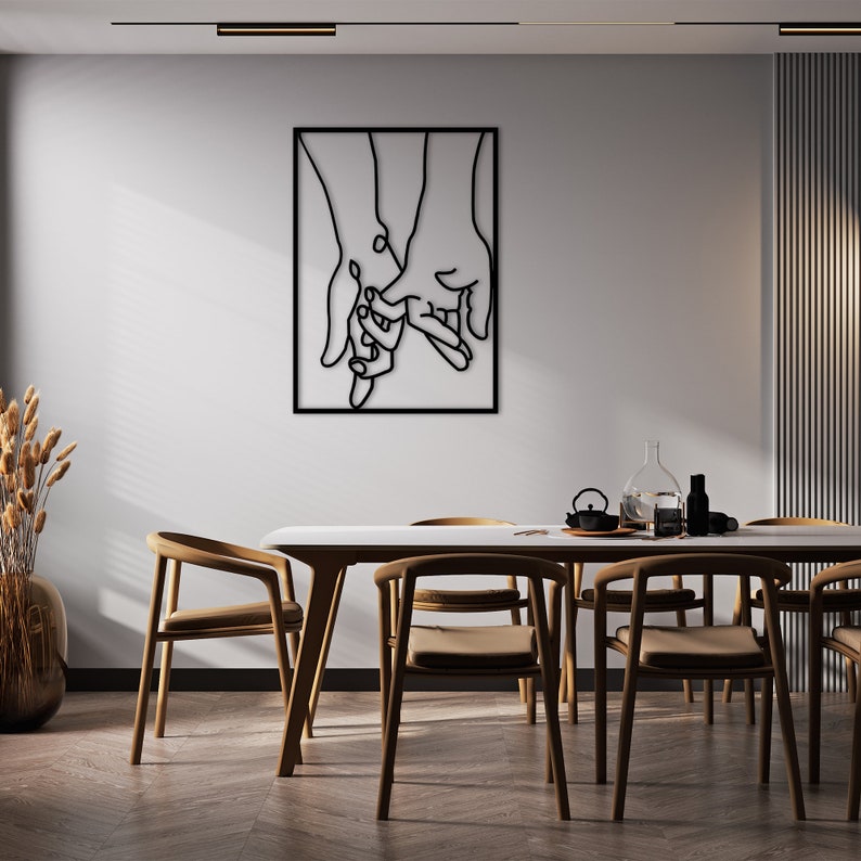 May include: Black metal wall art depicting two hands holding each other. The art is framed and hanging on a white wall above a wooden dining table with chairs.