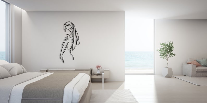 May include: A black line art wall decor of a person with a towel wrapped around their head, standing in a minimalist bedroom with a view of the ocean.