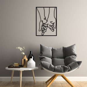 May include: Black metal wall art depicting two hands holding each other. The art is framed in a black square frame and is hanging on a gray wall.