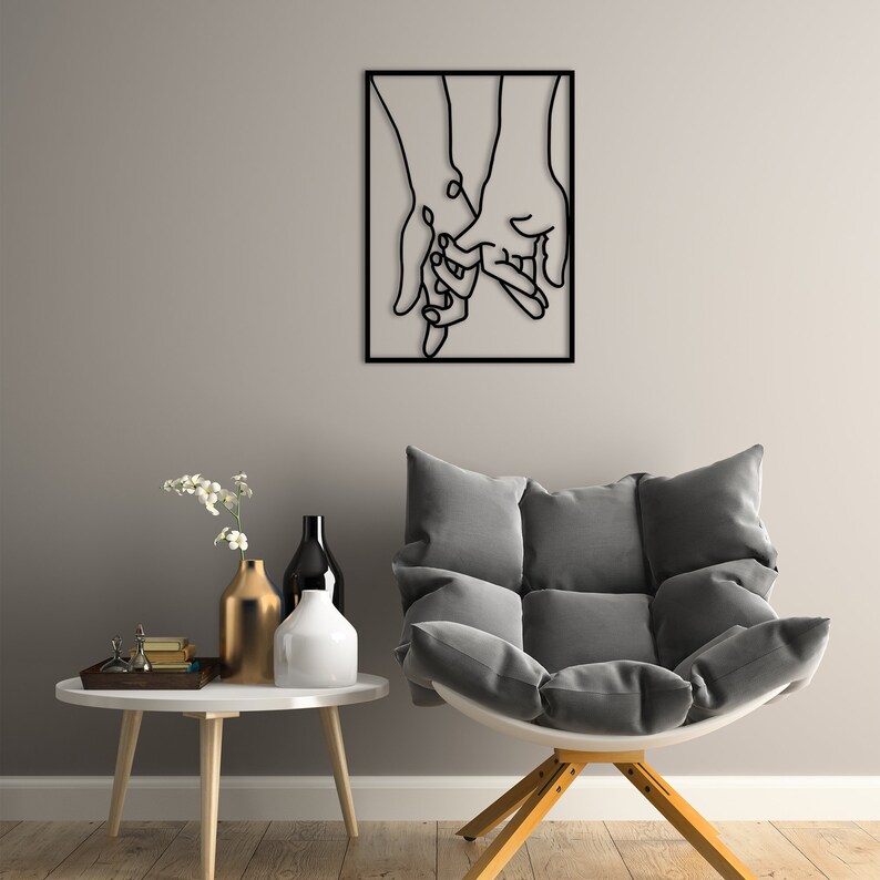 May include: Black metal wall art depicting two hands holding each other. The art is framed in a black square frame and is hanging on a gray wall.