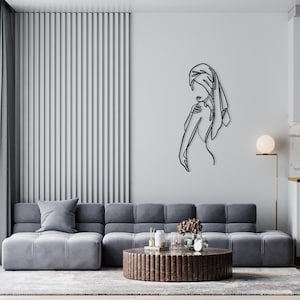 May include: A black metal wall sculpture of a woman with a towel wrapped around her head. The sculpture is on a white wall in a living room with a gray sofa and a brown coffee table.