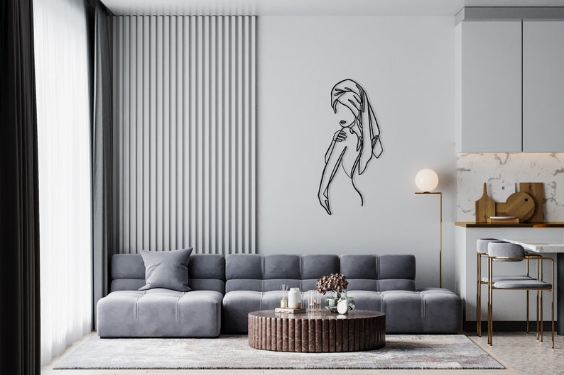 May include: A black metal wall sculpture of a woman with a towel wrapped around her head. The sculpture is on a white wall in a living room with a gray sofa and a brown coffee table.