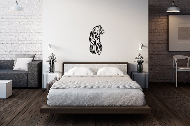 May include: A black metal wall art sculpture of a tiger in a modern bedroom setting. The sculpture is mounted on a white wall above a bed with white bedding and a dark wood frame. The bedroom has a white brick wall, a grey sofa, and a dark wood floor.