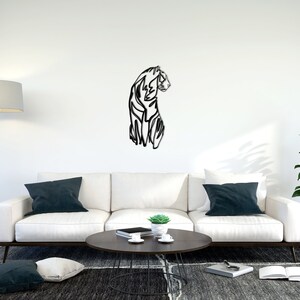 May include: A black and white line art metal wall sculpture of a tiger in a living room setting. The sculpture is mounted on a white wall above a white sofa with dark blue throw cushions. A coffee table with a book and a small potted plant sits in front of the sofa.