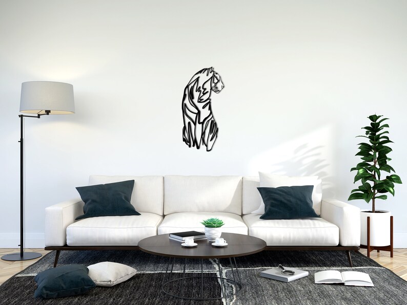 May include: A black and white line art metal wall sculpture of a tiger in a living room setting. The sculpture is mounted on a white wall above a white sofa with dark blue throw cushions. A coffee table with a book and a small potted plant sits in front of the sofa.