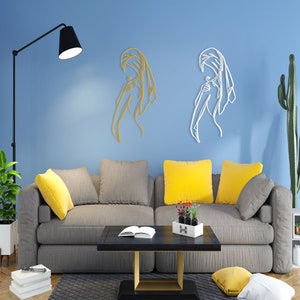 May include: Two metal wall sculptures of a woman in a flowing robe, one in gold and one in white, against a light blue wall. The sculptures are displayed above a gray sofa with yellow throw pillows.